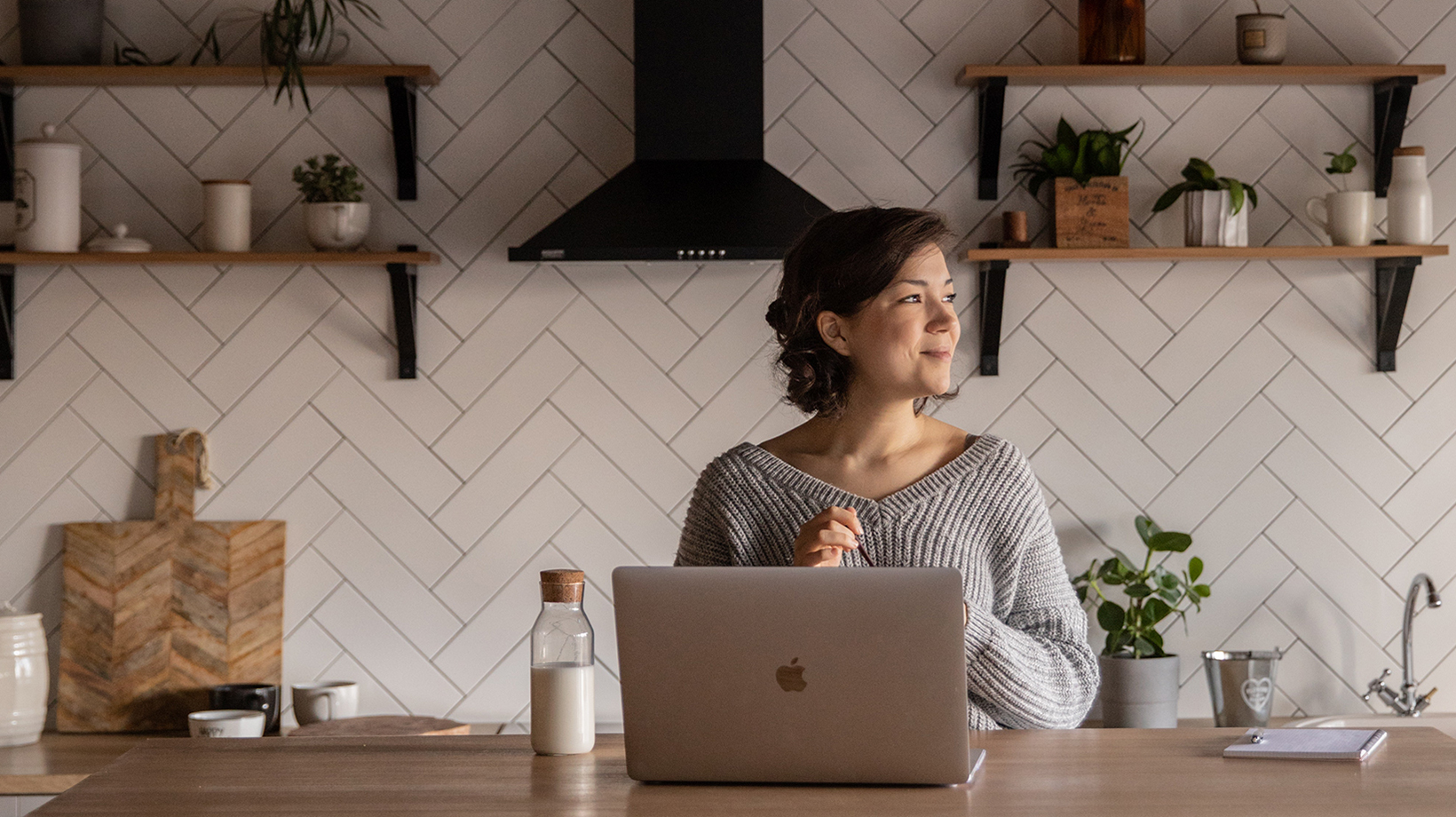 woman taking a break to look to what seems to be a window, while working on a laptop at a kitchen counter