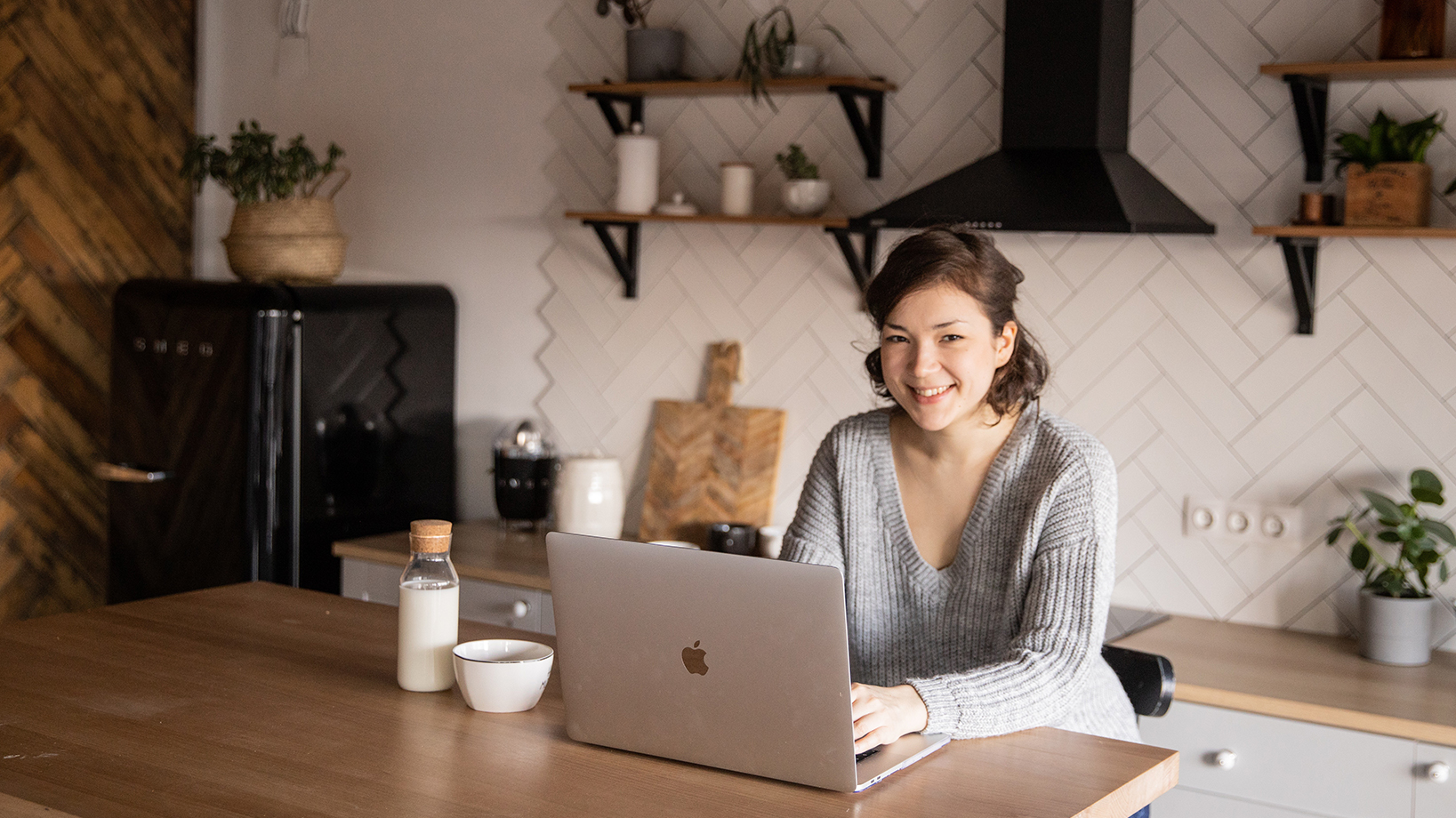 woman looking at the camera smiling while working on a laptop at a kitchen counter