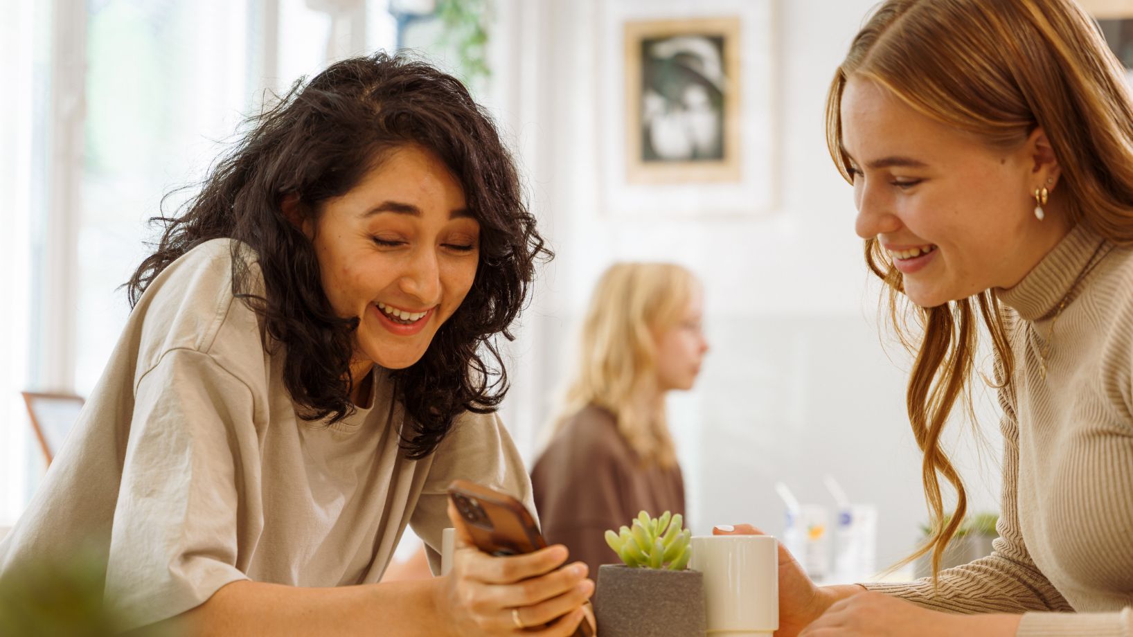 two women sitting at a table in a cafe, looking at something on a phone screen and laughing