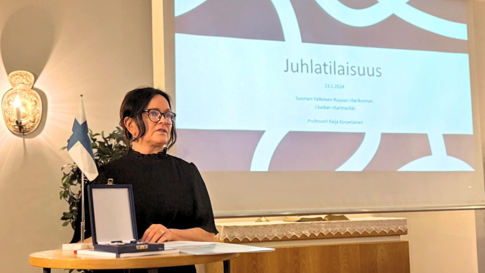 nainen puhujanpöydän takana Suomen lipun ja kunniamerkin kanssa